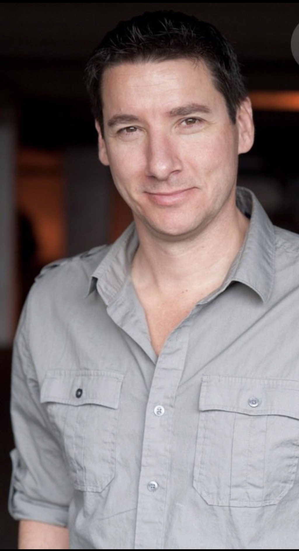 A man with short, dark hair and a slight smile is wearing a light gray button-up shirt